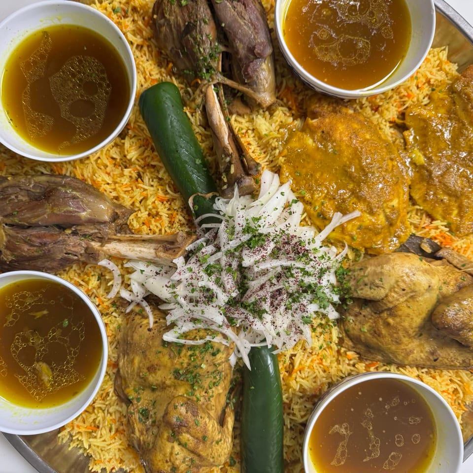 A large platter of Mediterranean rice with lamb shanks, chicken, sliced onions, and bowls of broth.